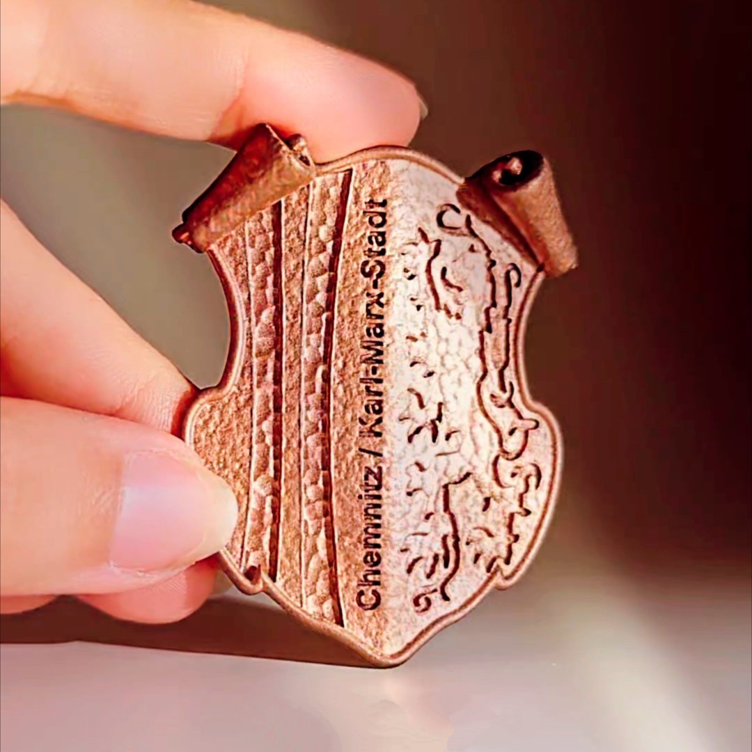 Bronzefarbener Magnet mit dem Stadtwappen von Chemnitz, gehalten zwischen zwei Fingern, inklusive Schriftzug „Chemnitz | Karl-Marx-Stadt“.