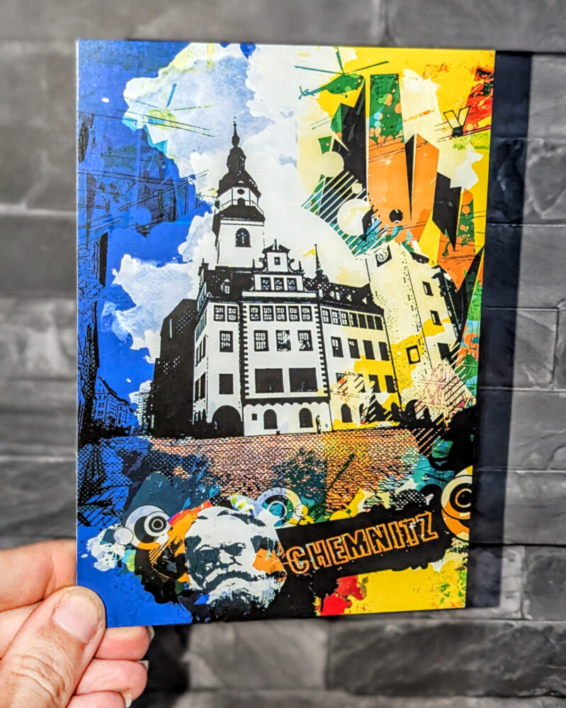 Bunte Pop-Art-Collage-Postkarte mit dem Alten Rathaus von Chemnitz, kombiniert mit grafischen Mustern, farbenfrohen Formen und einem Karl-Marx-Motiv. Die Karte wird von einer Hand vor einer grauen Steinwand gehalten.