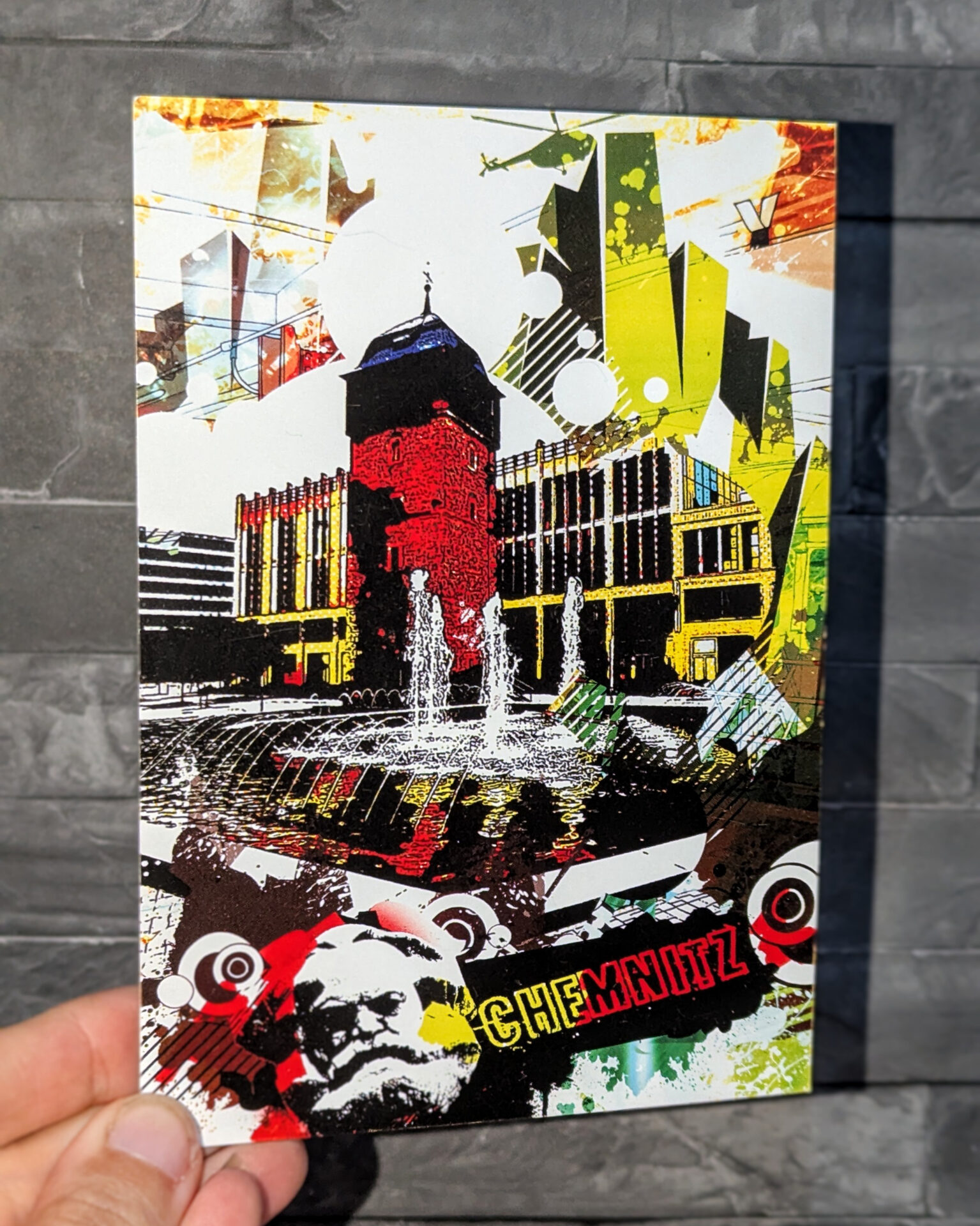 Bunte Collage-Postkarte mit dem Roten Turm in Chemnitz, dem Karl-Marx-Kopf und dem Brunnen am Markt, gehalten von einer Hand vor einer grauen Steinwand. Das Motiv zeigt farbenfrohe grafische Elemente und urbane Details im modernen Designstil.