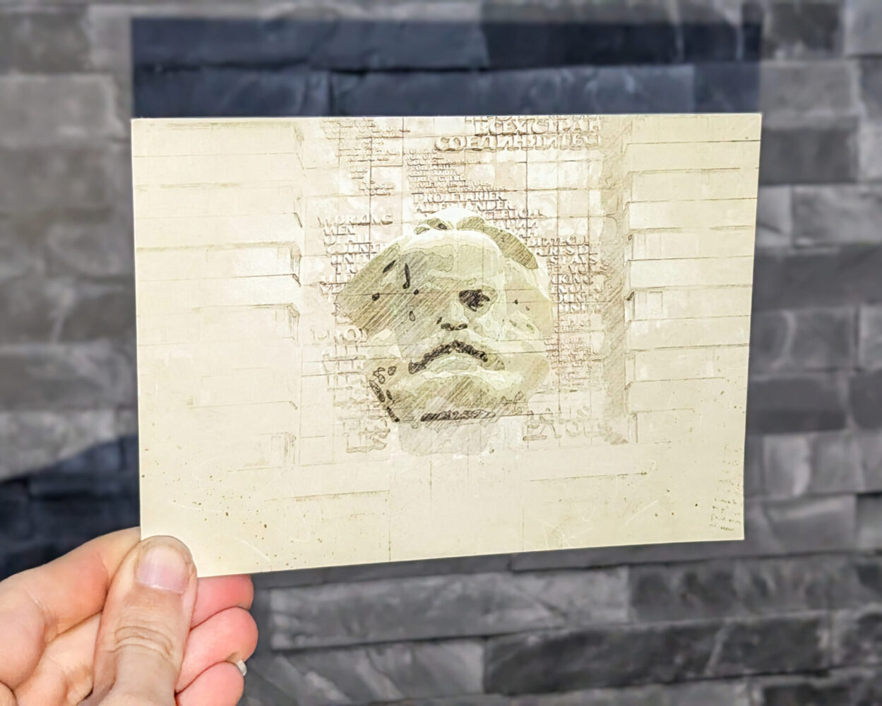 Künstlerische Postkarte mit einer gezeichneten Darstellung der Karl-Marx-Büste in Chemnitz, gehalten vor einer grauen Steinwand. Das Motiv wirkt im Vintage- und Skizzenstil.