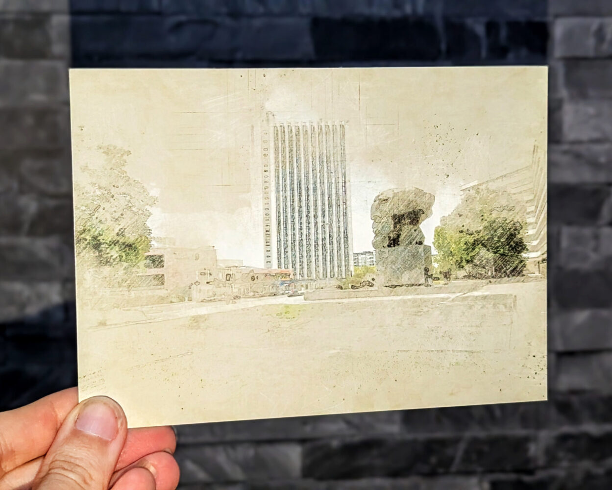 Künstlerische Postkarte mit einer gezeichneten Ansicht des Karl-Marx-Monuments in Chemnitz und dem angrenzenden Dorint Hotel, im weichen Sketch- und Vintage-Stil, gehalten vor einer grauen Steinwand.