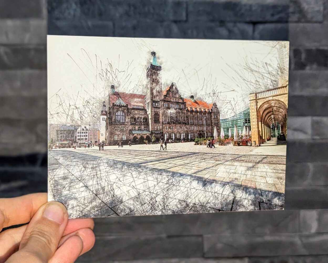 Künstlerische Postkarte vom historischen Rathaus in Chemnitz, im feinen Zeichenstil gestaltet. Die Karte wird von einer Hand vor einer grauen Steinwand gehalten und zeigt den Rathausplatz mit Menschen und umliegenden Gebäuden.