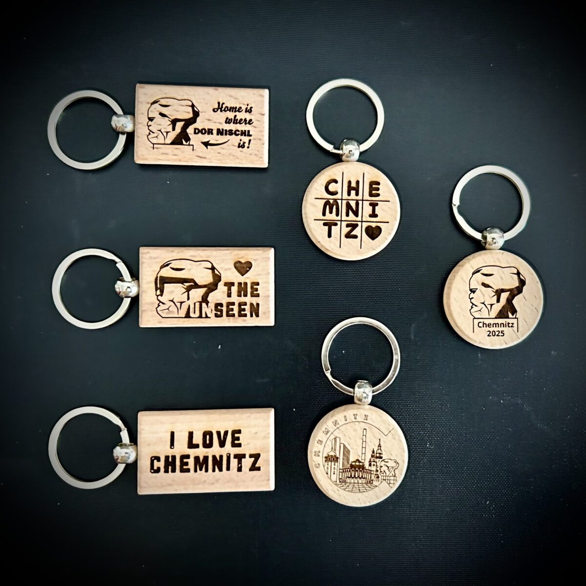 Schlüsselanhänger aus Holz mit verschiedenen Chemnitzer Motiven – darunter Karl-Marx-Kopf, Stadtsilhouette und typografische Designs; handgefertigte Geschenkidee aus Chemnitz.