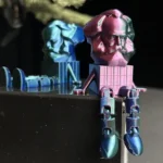3D-gedruckte Miniaturfiguren des Karl-Marx-Kopfes aus Chemnitz in metallic-blauen und pinken Farben, kreativ als sitzende Figuren gestaltet.